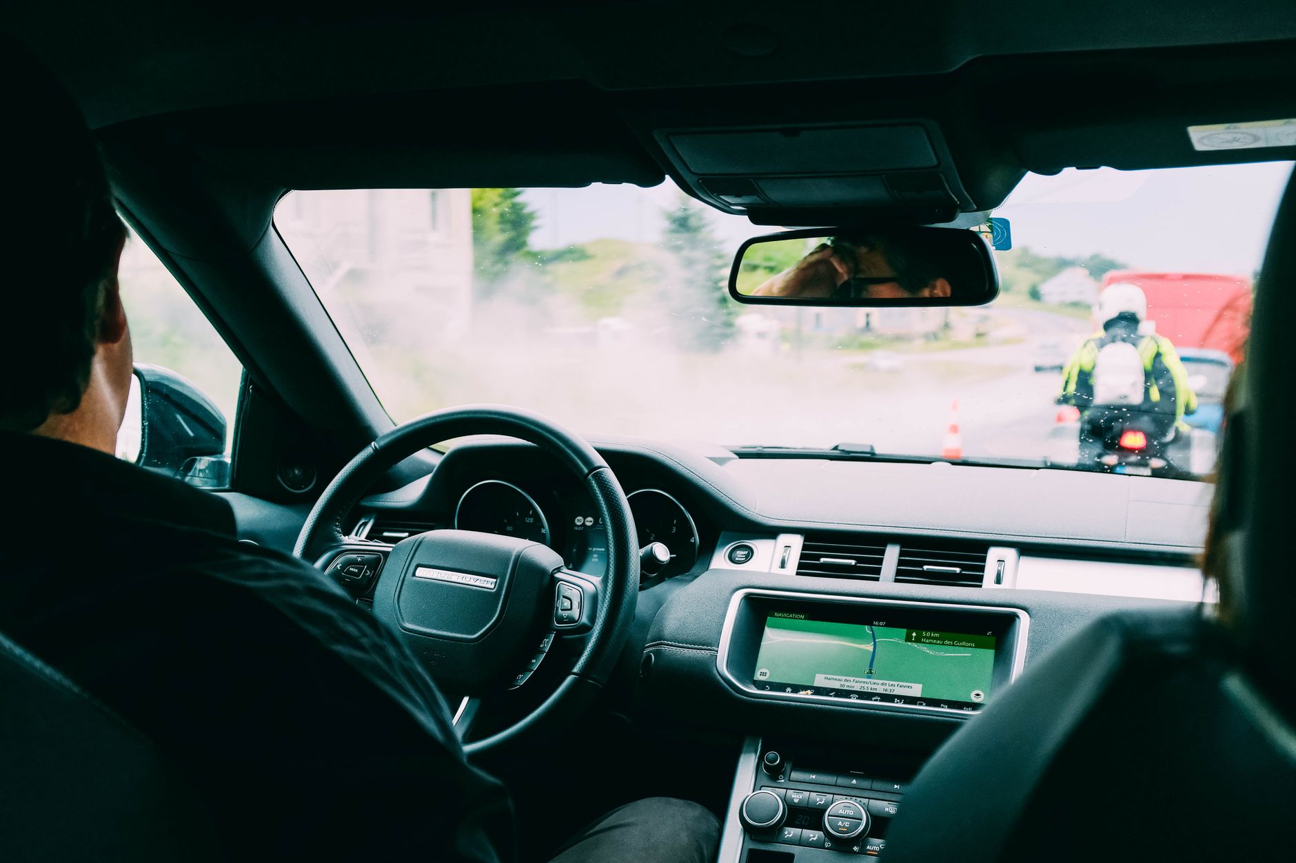 Intérieur d’une voiture vu de l’arrière : conducteur au volant, écran de navigation et motard en gilet fluorescent sur la route devant.