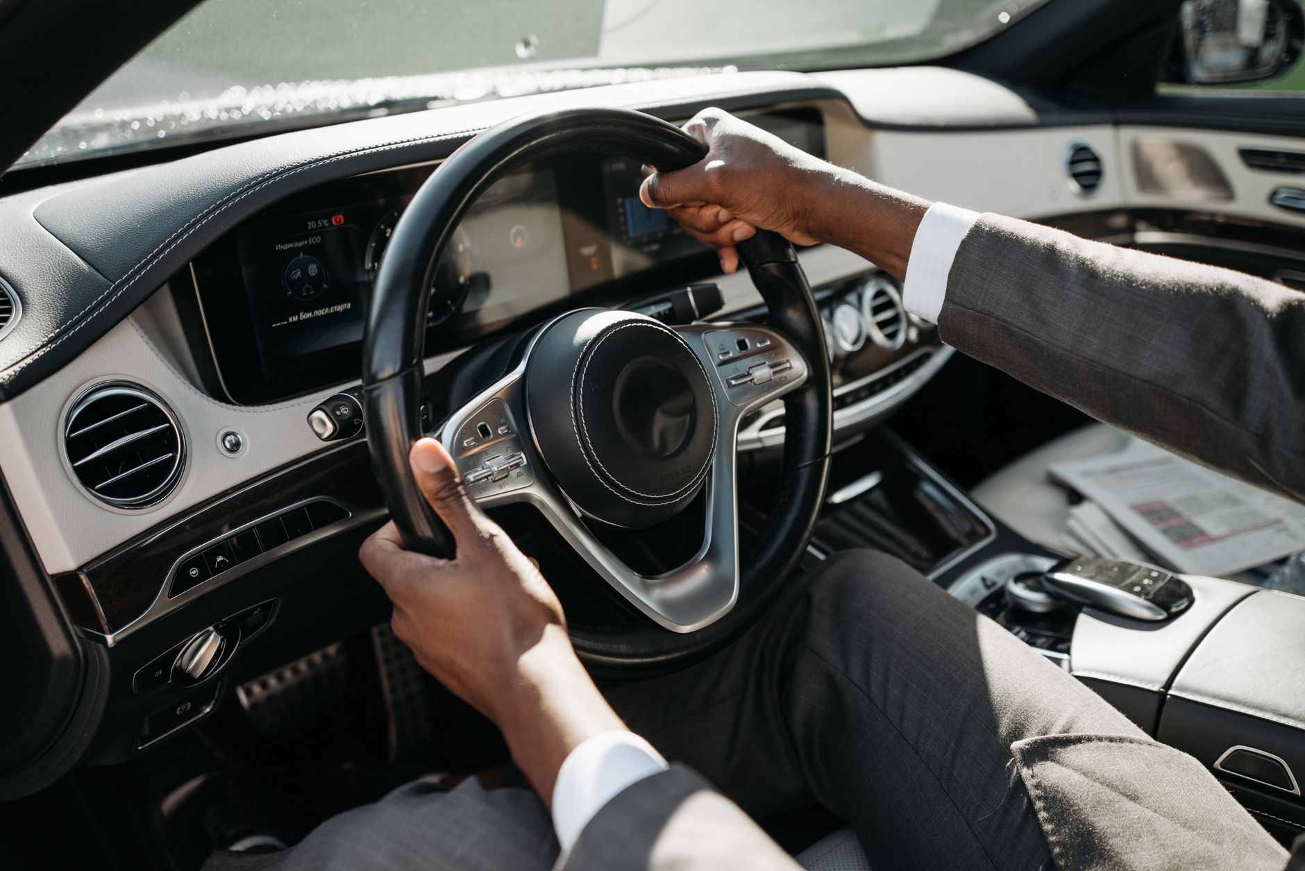 Mains d’une personne en costume gris sur le volant d’une voiture, avec le tableau de bord et la console centrale visibles.