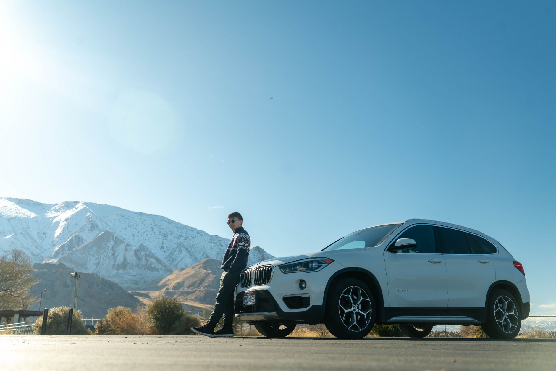 Une personne est appuyée contre l’avant d’un SUV blanc stationné devant un paysage de montagnes enneigées.